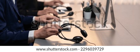 Panorama focus hand holding headset on call center workspace desk with blur background of operator team or telesales representative engaging in providing client with customer support service. Prodigy Royalty-Free Stock Photo #2296039729
