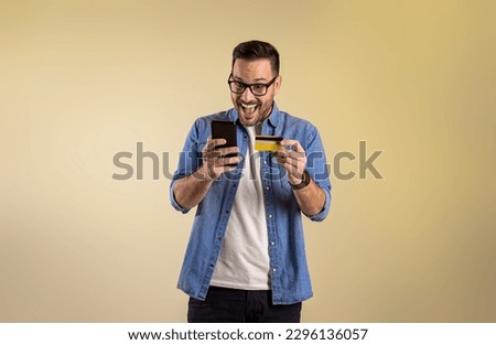 Male customer with credit card and smart phone screaming excitedly after winning rewards for online shopping. Cheerful young man buying through mobile apps on beige background Royalty-Free Stock Photo #2296136057