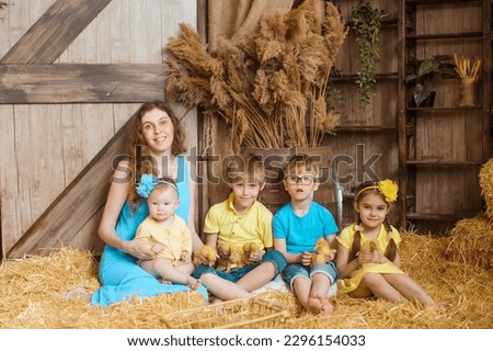 picture portrays a family of a mother and four children sitting on hay inside a barn. The children are holding ducklings and wearing summer clothes of yellow and blue. The mother has wavy hair.