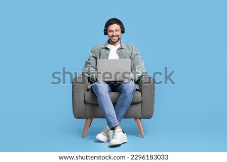 Happy man with laptop and headphones sitting in armchair on light blue background