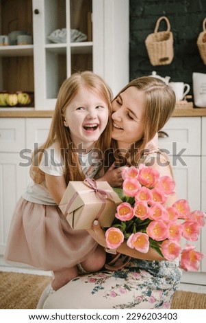 Daughter congratulates mother and gives present, bouquet of tulip flowers, in kitchen at home. Mother's day concept. Mom hug child girl smiling and surprised. Greeting card. International Women's Day. Royalty-Free Stock Photo #2296203403