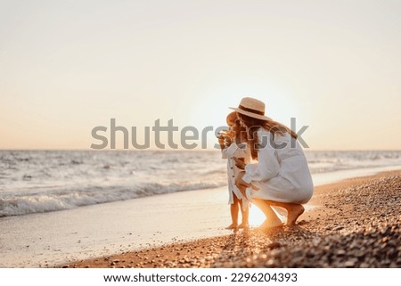 A young mother sat down and holds her little daughter by the hand on the ocean at sunset. Girls in white dresses and long hair.