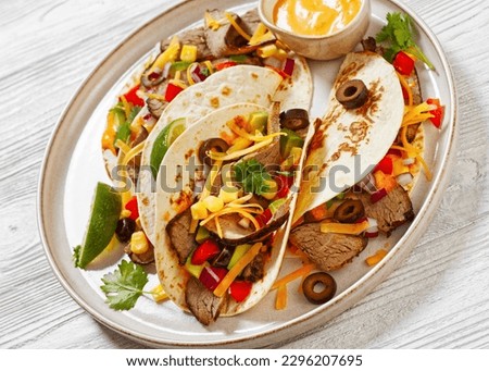 Grilled Steak Tacos with olives, tomatoes, red onion, avocado, cilantro, corn, shredded cheddar cheese and thousand island sauce on platter on white wood table, close-up