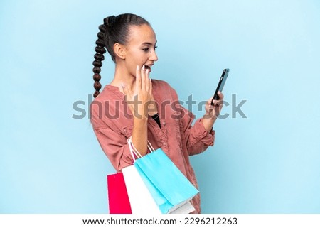 Young Arab woman isolated on blue background holding shopping bags and writing a message with her cell phone to a friend