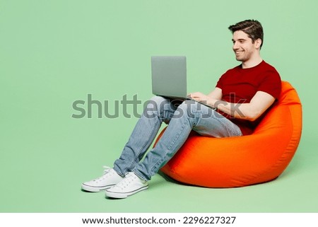 Full body smiling happy fun young IT man he wearing red t-shirt casual clothes sit in bag chair holding use work on laptop pc computer isolated on plain pastel light green background studio portrait
