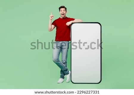 Full body young man he wearing red t-shirt casual clothes big huge blank screen mobile cell phone smartphone with workspace area point index finger up isolated on plain pastel light green background