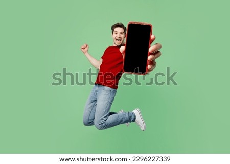 Full body young man wear red t-shirt casual clothes jump high hold in hand use close up mobile cell phone with blank screen workspace area do winner gesture isolated on plain pastel green background