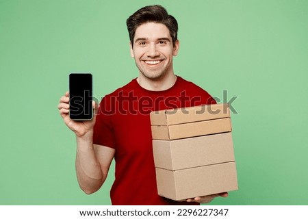 Young happy fun man he wears red t-shirt casual clothes hold stack cardboard blank boxes use mobile cell phone with blank screen workspace area isolated on plain pastel light green background studio