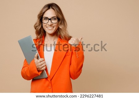 Young employee IT business woman corporate lawyer 30s wear classic formal orange suit glasses work in office hold closed laptop pc computer point finger aside isolated on plain beige background studio