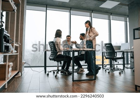 Against big windows by the table. Four people are working in the office together.