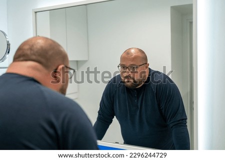 Confused upset overweight middle aged man looking at mirror in bathroom at home. Bearded male in eyeglasses dissatisfied with appearance. Midlife crisis, psychological problems, depression concept. Royalty-Free Stock Photo #2296244729