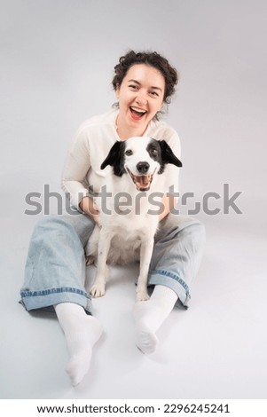 Happy laughing woman and her dog looking at camera sitting on grey floor. Owner young girl in blue jeans and white jumper having fun with her cute pet white young border collie  Royalty-Free Stock Photo #2296245241