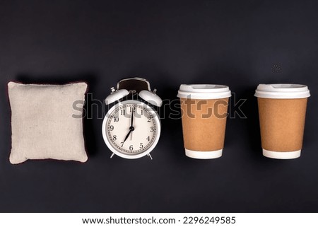 White alarm clock, disposable cups and a small pillow on a dark background.