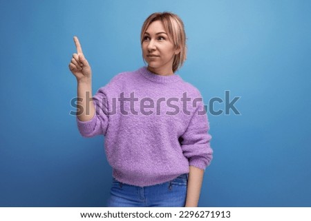 portrait of a cute charming pleasant blond young woman in a purple sweater pointing her finger to the side on a blue background with copy space