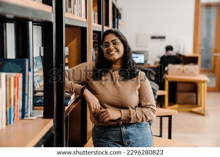 Young indian woman student in glasses smiling at camera while standing in college library