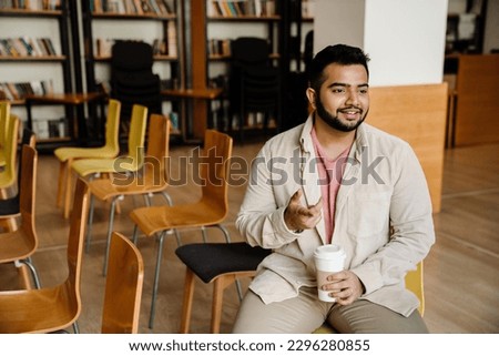 Young indian man student drinking coffee and talking to somebody while sitting in college library