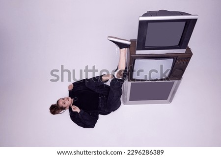 Top view. Beautiful young girl in jeans clothes lying on floor near retro tv sets over grey background. News, information. Concept of fashion, 80s, 90s style, retro and vintage, gadgets, mass media Royalty-Free Stock Photo #2296286389