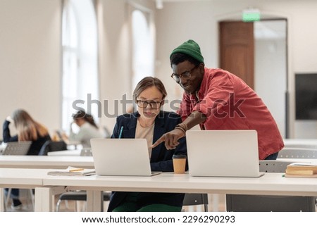 Smiling african american guy helps caucasian girl students working on project in college university library. Positive man woman look at laptop screen explaining studying learning together in campus.