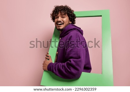 Happy Indian man looking out of the picture frame while standing against pink background
