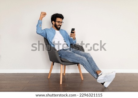 Great News. Joyful Young Indian Man Looking At Smartphone Screen And Celebrating Success While Sitting In Chair At Home, Happy Eastern Male Exclaiming With Excitement And Shaking Fist, Copy Space Royalty-Free Stock Photo #2296321115