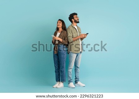 Dating mobile app concept. Stylish young eastern man and woman hipsters standing back to back, using smartphones on colorful studio background, chatting online, looking at copy space, full length