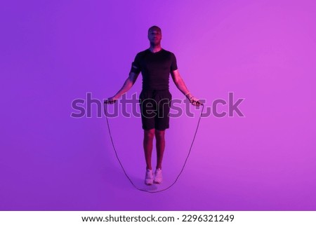 Black Sportsman Exercising With Jumping Rope Having Fitness Workout Posing Over Purple Neon Studio Background. Full Length Shot Of Sporty Man Training Doing Jumps Looking At Camera