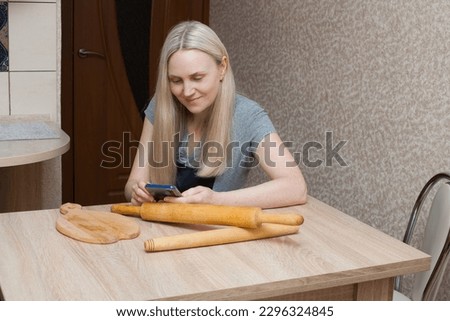 Smiling blonde woman in a T-shirt and apron talking on the phone at a table in the kitchen. On the table are wooden tools for cooking. A live shot of a woman's everyday life at home.