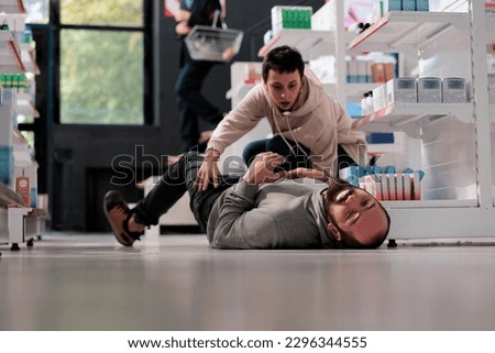 Terrified woman noticing man on drugstore floor having seizure, running to give first aid for fainting. Apothecary client helping person suffering from epilepsy disorder attack Royalty-Free Stock Photo #2296344555