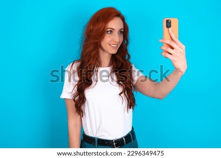 Isolated shot of pleased cheerful Young redhead woman wearing white T-shirt over blue background , makes selfie with mobile phone. People, technology and leisure concept