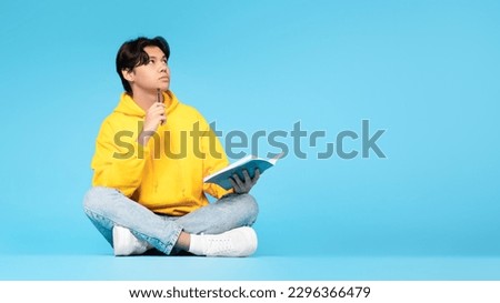 Pensive Asian Student Guy Thinking Holding Textbook And Pen Learning Sitting On Floor Over Blue Studio Background. Thoughtful Teen Boy Doing His Homework Taking Notes. Panorama With Free Space Royalty-Free Stock Photo #2296366479