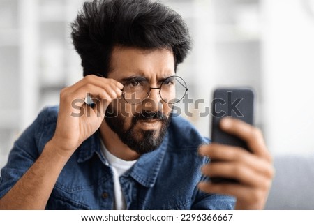 Eyesight Problems Concept. Young Indian Man In Eyeglasses Looking At Smartphone Screen And Squinting, Eastern Guy Trying To Read Message, Suffering From Astigmatism And Bad Vision, Closeup Shot Royalty-Free Stock Photo #2296366667