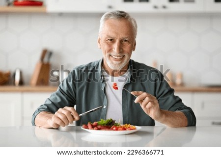Handsome Smiling Senior Man Eating Tasty Meal In Kitchen At Home, Happy Elderly Gentleman Sitting At Table And Looking At Camera, Enjoying Delicious Food, Holding Fork And Knife, Copy Space Royalty-Free Stock Photo #2296366771