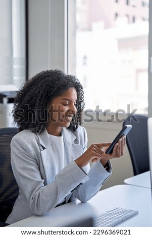 Vertical portrait of female entrepreneur businesswoman, successful African American business woman using smartphone, cellphone application, online communication, sitting at table in office indoors.
