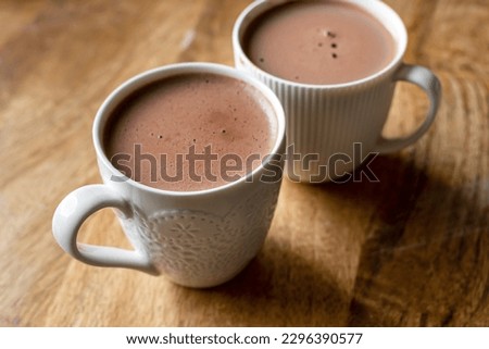 White mugs filled with rich, steaming cocoa on a rustic wooden table. The cozy and warm ambiance, coupled with the visual appeal of the white mugs and wooden texture, visual for coffee shops or cafes