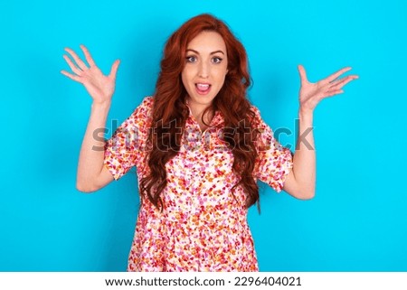 Young redhead woman wearing floral dress over blue background raising hands up, having eyes full of happiness rejoicing his great achievements. Achievement, success concept.