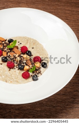 Oatmeal with nuts of various kinds, raspberries and blueberries with mint leaves in a light ceramic plate. The dish stands on a wooden table, next to a glass with orange juice.