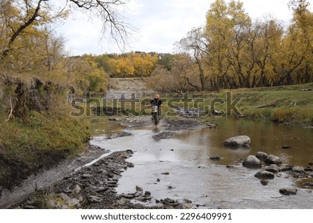 Dirt bike rider racing through river bed with autumn scenery in background