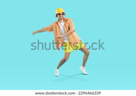 Happy funny tourist in summer clothes dancing isolated on blue background. Portrait of handsome young man wearing orange holiday shirt, yellow shorts, bucket hat and sun glasses dancing and having fun