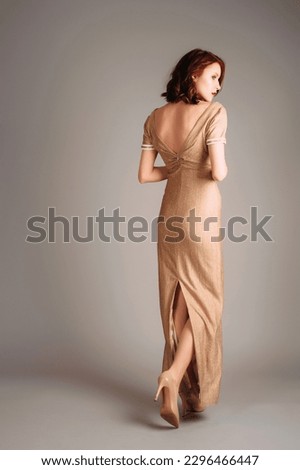Ivory maxi dress with gold glitter, short sleeves and v line neckline. Beautiful tall red-haired young woman walking in slim backless evening gown and high heels. Total beige look. Rear view. Royalty-Free Stock Photo #2296466447
