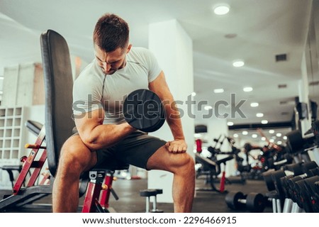 A strong muscular sportsman is sitting in a gym during his strength training and pumping muscles while looking at it. A dedicated sportsman is sitting in a gym and doing exercises for the biceps. Royalty-Free Stock Photo #2296466915