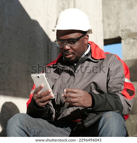 African american worker in hard hat and protective goggles with smartphone sits at construction site. Workman holds in hands white smartphone and talks by video link