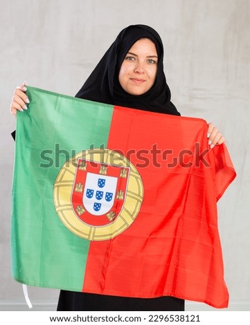 joyful smiling Muslim woman in traditional black hijab holds flag of Portugal. Portrait of female Muslim with Portuguese flag on gray background