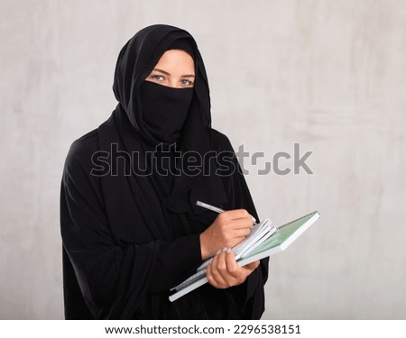 young calm girl with covered face by black burka holds several notebooks in her hands and writes important note in her diary