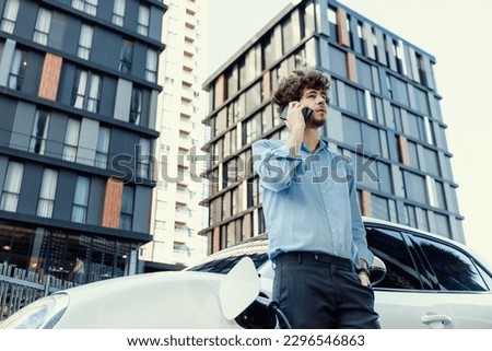 Progressive businessman talking on the phone, leaning on electric car recharging with public EV charging station, apartment condo residential building on the background as green city lifestyle.