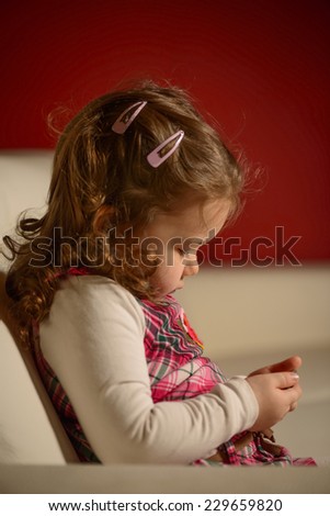 Pretty little girl with brown long hair, dressed with pink striped dress, sitting on sofa in living room,  holding mobile phone watching cartoons