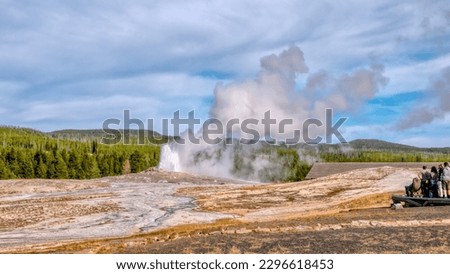 At Yellowstone National Park, Old Faithful beginning to erupt with water and steam rising, as unidentifiable tourists watch from a viewing platform on the side. Royalty-Free Stock Photo #2296618453