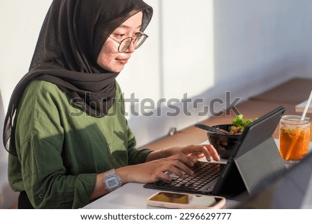 young woman in hijab sits working in a cafe using digital tablet and keyboard, next to her are drinks and healthy food