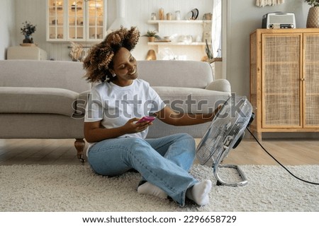Happy relaxed young African American woman holding smartphone sitting in front of electric fan at home, smiling black girl feeling good while relaxing near ventilator during extreme heat Royalty-Free Stock Photo #2296658729