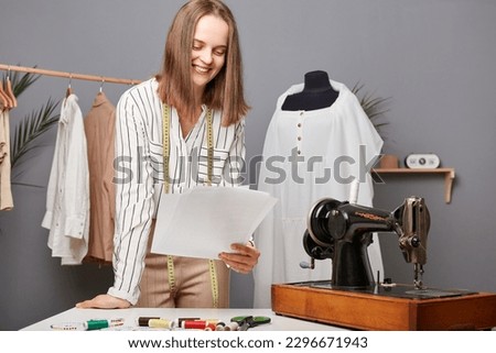Small garment business owner. Caucasian fashion designer holding papers with sketch in workshop, thinking about new ides for dresses, needlewoman working process.