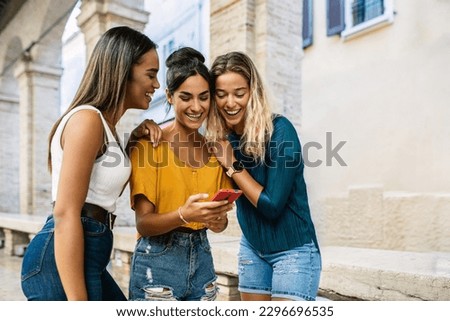 Three diverse young women having fun using mobile phone together in city street. Technology lifestyle and social media concept. Royalty-Free Stock Photo #2296696535